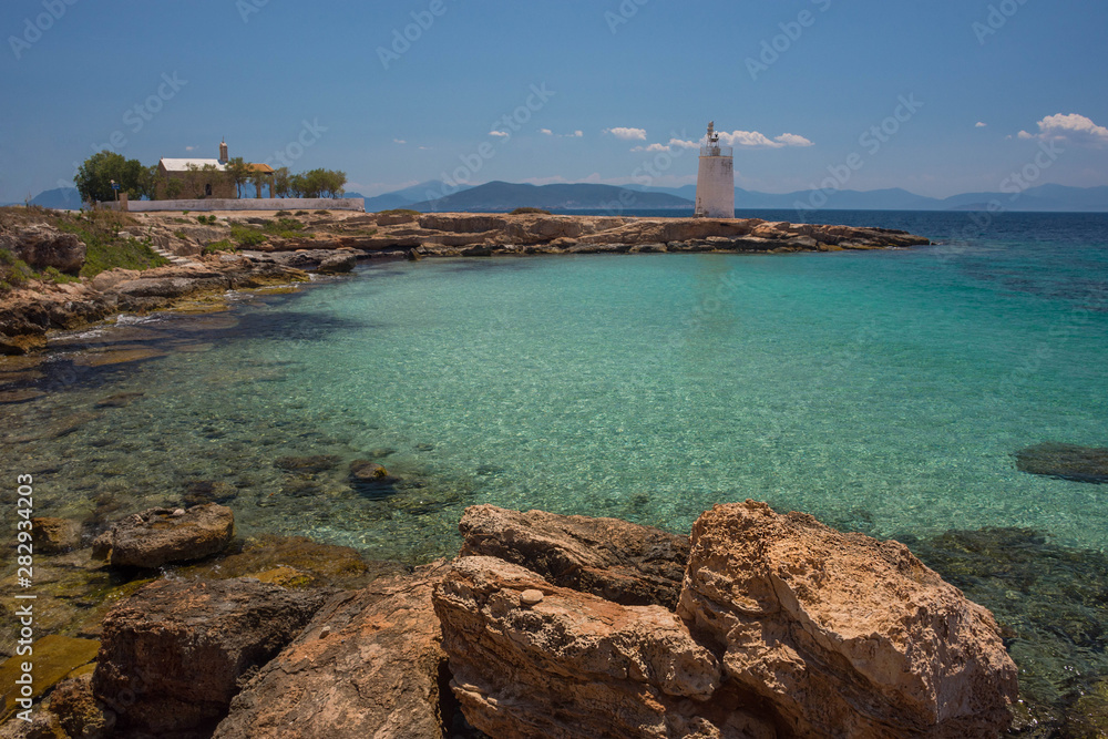 The wild coast of Aegina island with clear and blue waters of Mediterranean sea and the old small lighthouse in the background, in Saronic gulf, Greece.