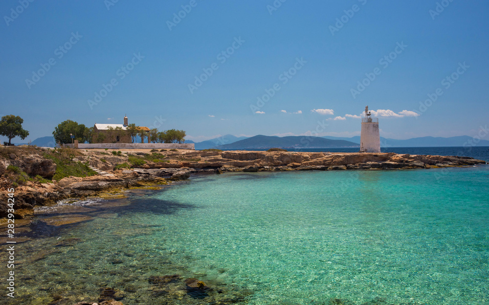 The wild coast of Aegina island with clear and blue waters of Mediterranean sea and the old small lighthouse in the background, in Saronic gulf, Greece.