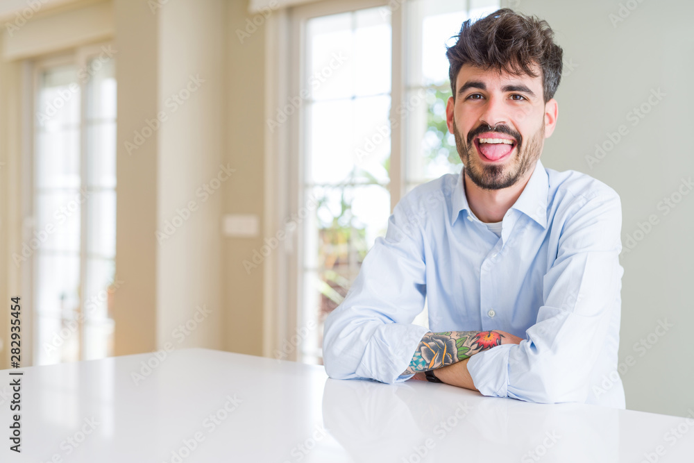 Fototapeta premium Young businesss man sitting on white table sticking tongue out happy with funny expression. Emotion concept.