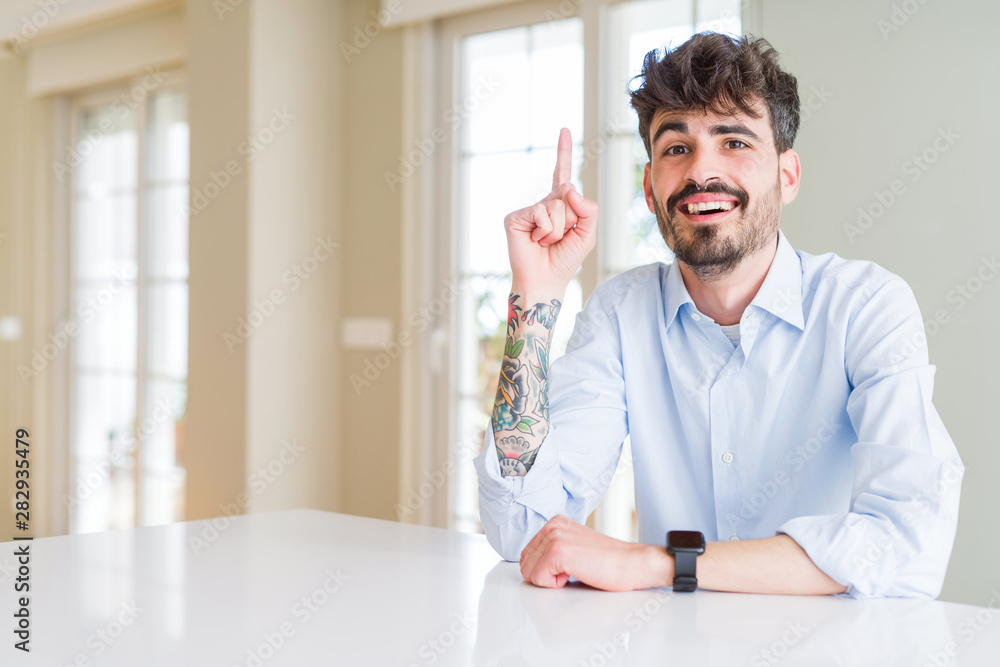 Fototapeta premium Young businesss man sitting on white table pointing finger up with successful idea. Exited and happy. Number one.