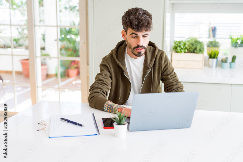 Young student man using computer laptop and notebook scared in shock with a surprise face, afraid and excited with fear expression Young student man using computer laptop and notebook scared in shock with a surprise face, afraid and excited with fear expression