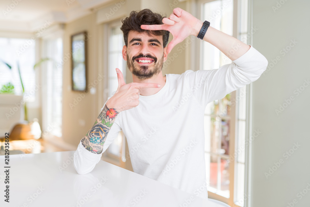 Young man wearing casual shirt sitting on white table smiling making frame with hands and fingers with happy face. Creativity and photography concept. Young man wearing casual shirt sitting on white table smiling making frame with hands and fingers with happy face. Creativity and photography concept.