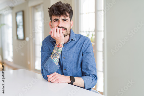 Young man wearing casual shirt sitting on white table looking stressed and nervous with hands on mouth biting nails. Anxiety problem.