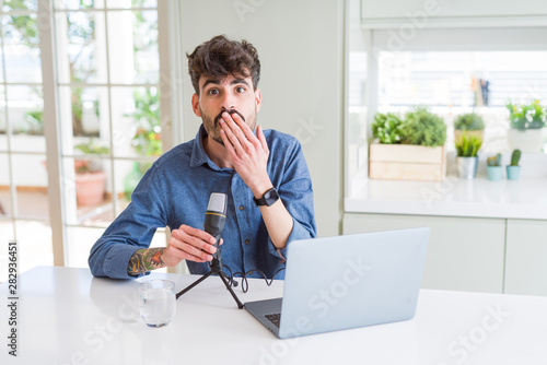 Young man recording podcast using microphone and laptop cover mouth with hand shocked with shame for mistake, expression of fear, scared in silence, secret concept