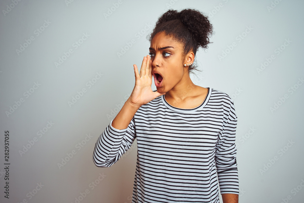 African american woman wearing navy striped t-shirt standing over isolated white background shouting and screaming loud to side with hand on mouth. Communication concept. African american woman wearing navy striped t-shirt standing over isolated white background shouting and screaming loud to side with hand on mouth. Communication concept.