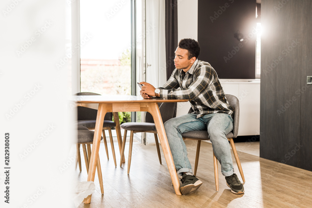 full length view of handsome african american man sitting before table and using smartphone