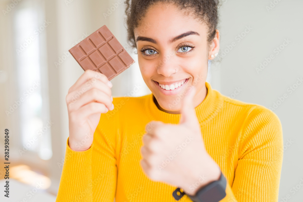 Young african american girl eating dark chocolate bar as a sweet snack happy with big smile doing ok sign, thumb up with fingers, excellent sign Young african american girl eating dark chocolate bar as a sweet snack happy with big smile doing ok sign, thumb up with fingers, excellent sign
