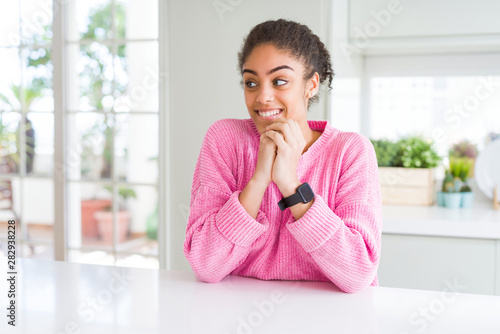 Beautiful african american woman with afro hair wearing casual pink sweater laughing nervous and excited with hands on chin looking to the side