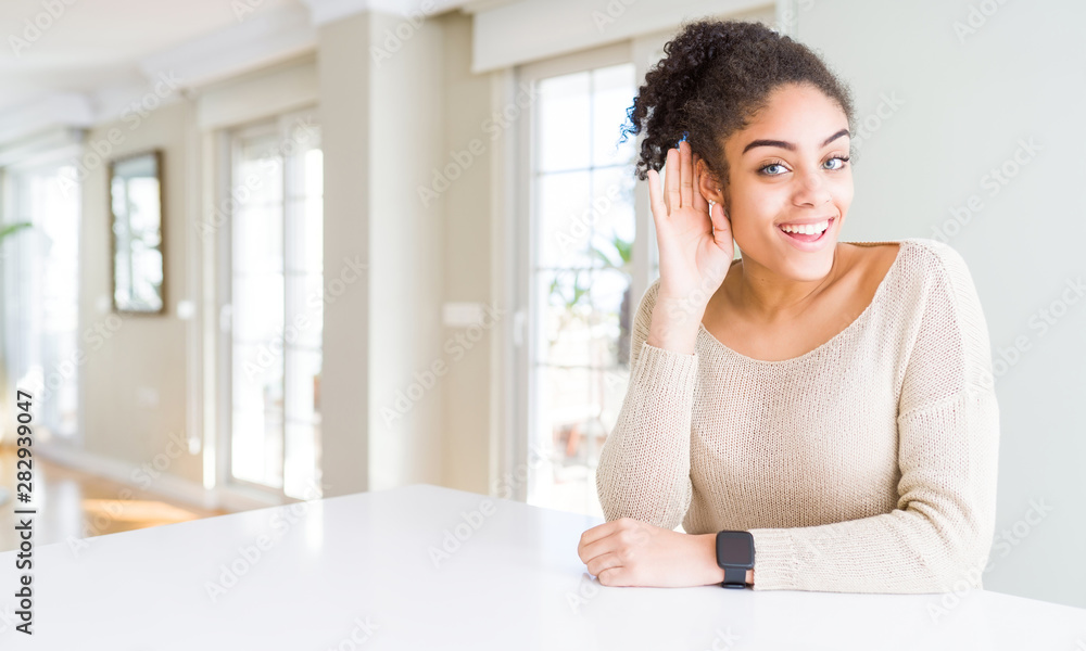 Beautiful young african american woman with afro hair sitting on table at home smiling with hand over ear listening an hearing to rumor or gossip. Deafness concept. Beautiful young african american woman with afro hair sitting on table at home smiling with hand over ear listening an hearing to rumor or gossip. Deafness concept.