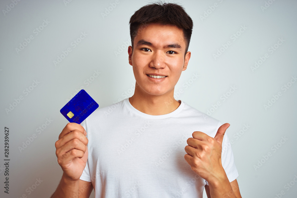 Young asian chinese man holding credit card standing over isolated white background happy with big smile doing ok sign, thumb up with fingers, excellent sign Young asian chinese man holding credit card standing over isolated white background happy with big smile doing ok sign, thumb up with fingers, excellent sign