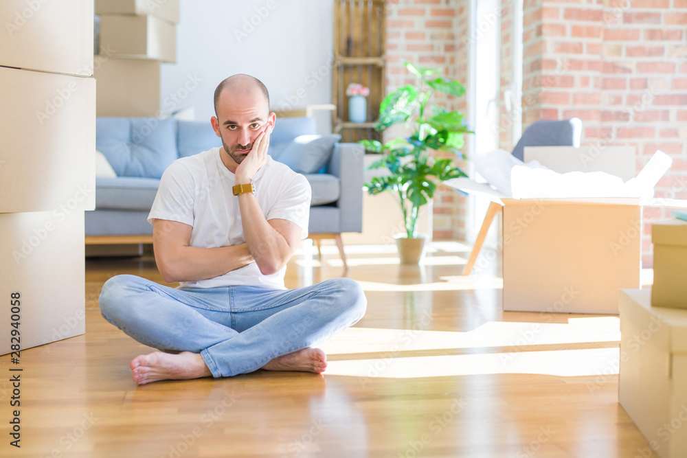 Young bald man sitting on the floor around cardboard boxes moving to a new home thinking looking tired and bored with depression problems with crossed arms. Young bald man sitting on the floor around cardboard boxes moving to a new home thinking looking tired and bored with depression problems with crossed arms.