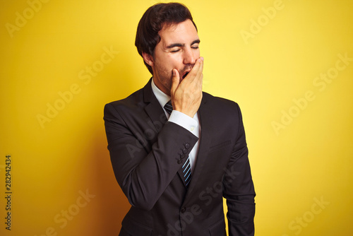 Young handsome businessman wearing suit and tie standing over isolated yellow background bored yawning tired covering mouth with hand. Restless and sleepiness.