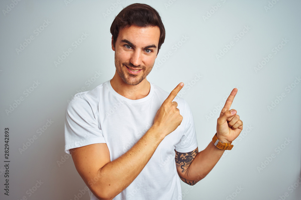 Young handsome man with tattoo wearing casual t-shirt over isolated white background smiling and looking at the camera pointing with two hands and fingers to the side. Young handsome man with tattoo wearing casual t-shirt over isolated white background smiling and looking at the camera pointing with two hands and fingers to the side.