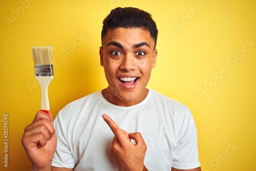 Young brazilian painter man holding brush standing over isolated yellow background very happy pointing with hand and finger