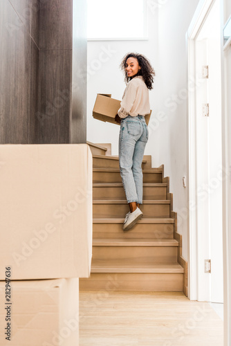 full length view of beautiful african american woman walking upstairs while holding box and looking at camera