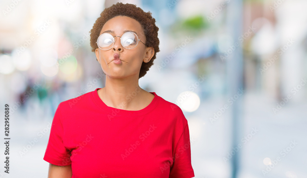 Beautiful young african american woman wearing glasses over isolated background making fish face with lips, crazy and comical gesture. Funny expression.