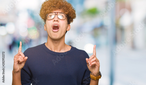 Young handsome man with afro wearing glasses amazed and surprised looking up and pointing with fingers and raised arms.