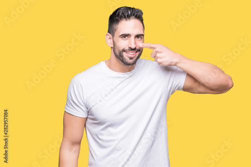 Handsome man wearing white t-shirt over yellow isolated background Pointing with hand finger to face and nose, smiling cheerful