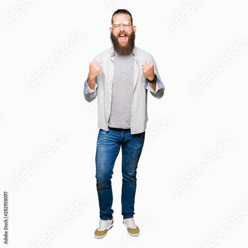 Young blond man wearing glasses very happy and excited doing winner gesture with arms raised, smiling and screaming for success. Celebration concept.