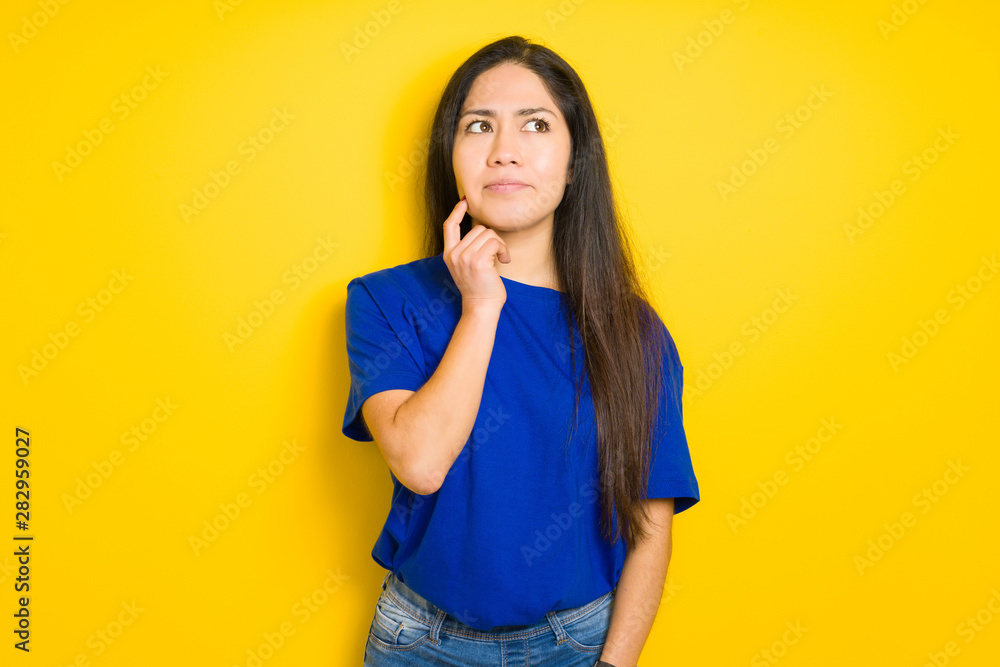 Beautiful brunette woman wearing blue t-shirt over yellow isolated background with hand on chin thinking about question, pensive expression. Smiling with thoughtful face. Doubt concept. Beautiful brunette woman wearing blue t-shirt over yellow isolated background with hand on chin thinking about question, pensive expression. Smiling with thoughtful face. Doubt concept.