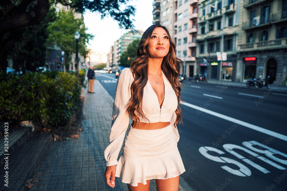 Fototapeta premium Summer sunny lifestyle portrait of young stylish asian girl on the street of big modern city. She wears white dress. women business