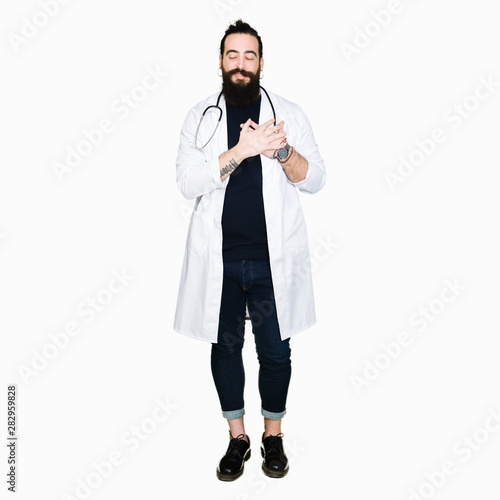 Doctor with long hair wearing medical coat and stethoscope smiling with hands on chest with closed eyes and grateful gesture on face. Health concept.