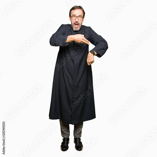 Middle age priest man wearing catholic robe In hurry pointing to watch time, impatience, upset and angry for deadline delay