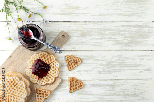 Still life with homemade cakes and blueberry jam. Sweet waffles on a wooden Board with a jar of jam and a bouquet of daisies on a wooden background. The concept of morning Breakfast and home comfort