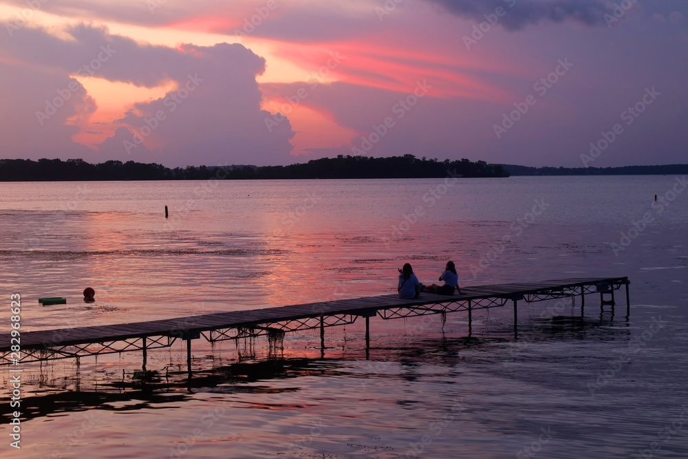 Beautiful summer landscape with colorful after sunset sky.Scenic view with bright color sky reflects in a lake and two silhouettes on a wooden pier enjoying sunset over lake Mendota. Madison, WI, USA.