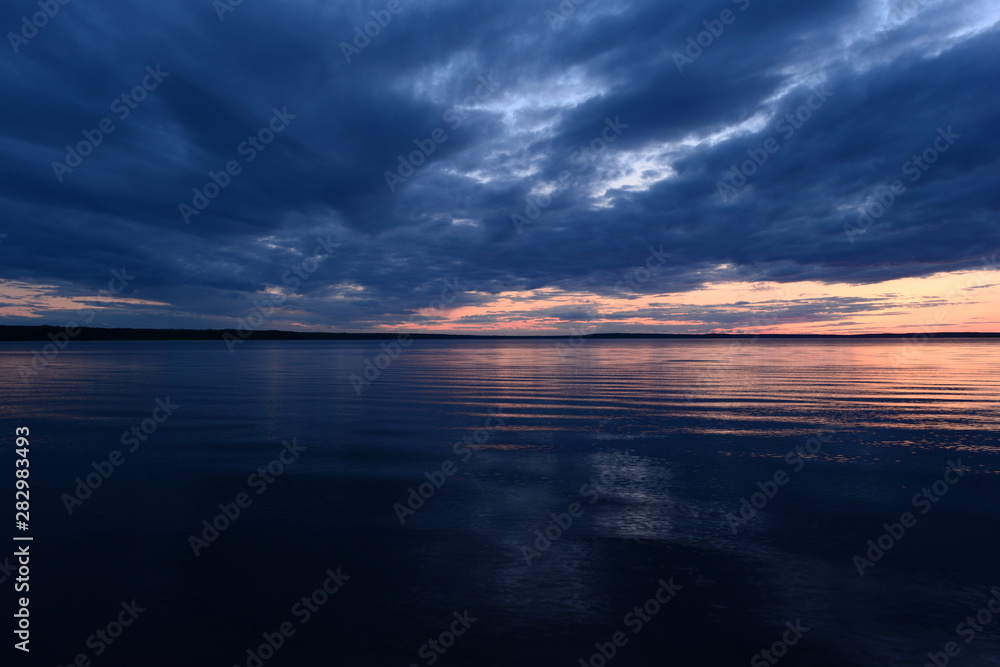 Twilight sky in bright blue clouds above a quiet surface of the water in the light of the setting sun before night