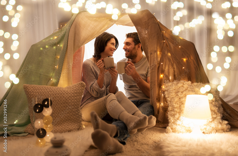 leisure, hygge and people concept - happy couple drinking coffee or tea in kids tent at home