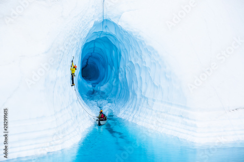 Ice cimber in front of an ice cave leading up from a canoe in a blue pool on the Matanuska Glacier.