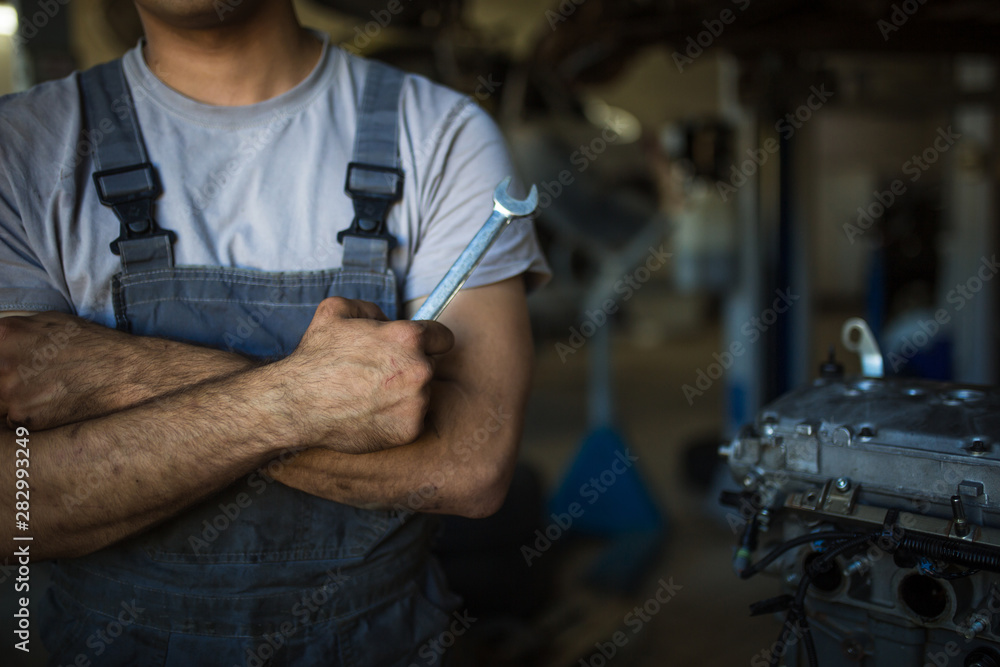A mechanic in overalls with tools in his hands stands with his arms crossed on his chest. Work in the service station in the background. A mechanic in overalls with tools in his hands stands with his arms crossed on his chest. Work in the service station in the background.