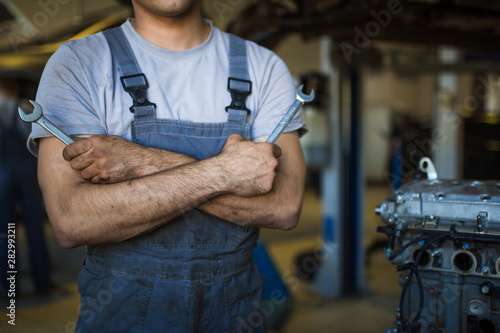 A mechanic in overalls with tools in his hands stands with his arms crossed on his chest. Work in the service station in the background.