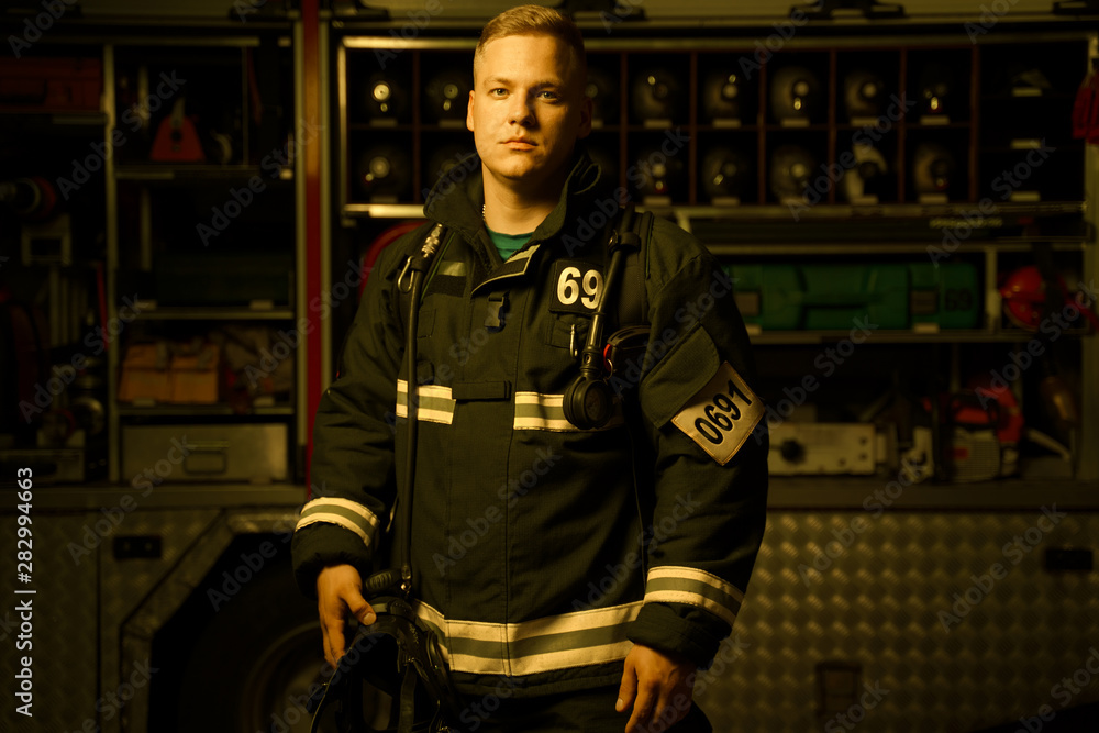 Full-length image of young firefighter man with helmet in his hand looking into camera while Full-length image of young firefighter man with helmet in his hand looking into camera while