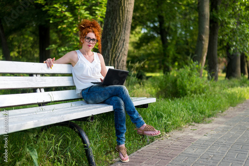 Red-haired female freelancer working on a laptop while sitting on a park bench.