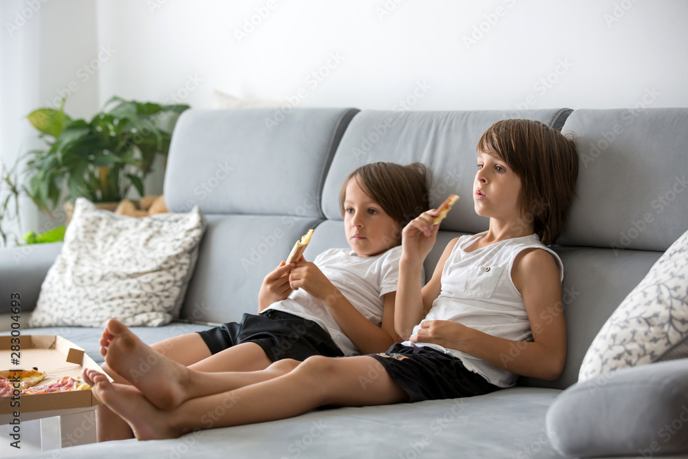 Cute children, sitting on couch, eating pizza and watching TV. Hungry child taking a bite from pizza on a pizza party Cute children, sitting on couch, eating pizza and watching TV. Hungry child taking a bite from pizza on a pizza party