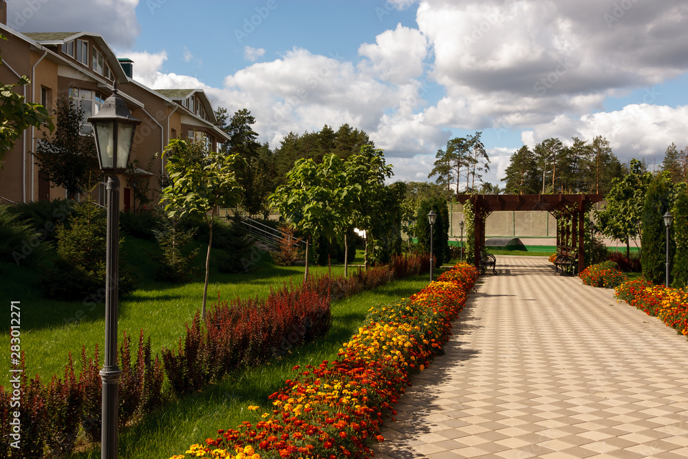 Fototapeta premium A quiet street in a modern country village on a sunny summer day. In the foreground Street lamp. The paved path is laid past flower beds, ornamental shrubs and trees.