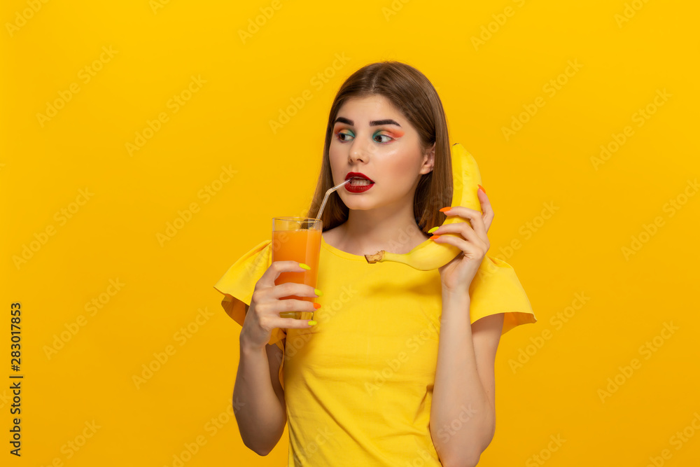 Close-up portrait of a beautiful young girl holding glass of ofange juice and banana close to face isolated over yellow background. Close-up portrait of a beautiful young girl holding glass of ofange juice and banana close to face isolated over yellow background.