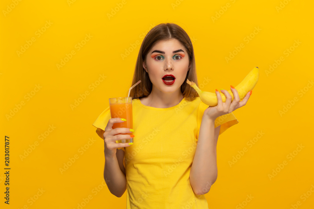 Fototapeta Close-up portret pięknej młodej dziewczyny trzymającej szklankę soku orange i banana blisko twarzy na białym tle na żółtym tle.