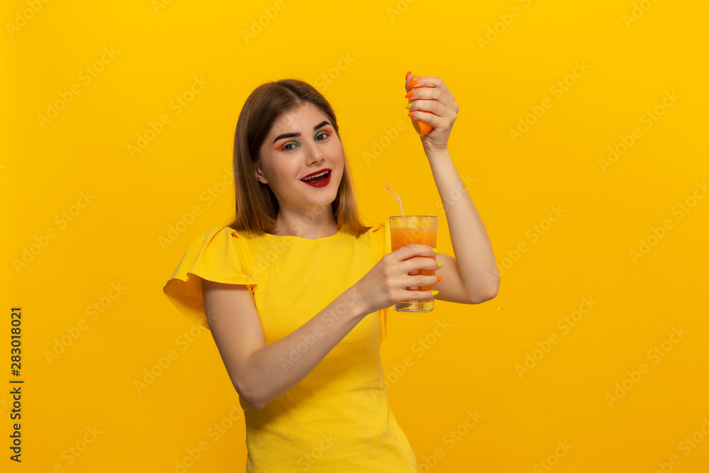 Close-up portrait of a beautiful young girl squashing orange juice into glass isolated over yellow background. Close-up portrait of a beautiful young girl squashing orange juice into glass isolated over yellow background.