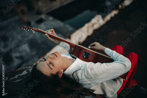 A young woman lies and plays guitar on an old abandoned ship in white shirt and red tights.