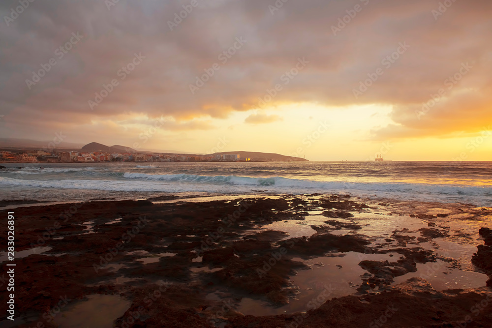 Tropical sunrise over the limestone coasts of El Medano, Tenerife, Canary Islands, Spain, beautiful seascape with vibrant colors overcast sky, golden haze light and dynamic waves striking rocky shores