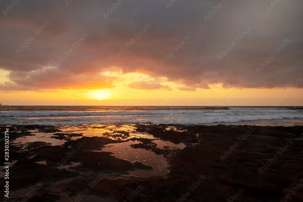 Glorious sunrise reflected in the natural rock pools formed in the limestone shores of El Medano resort, Tenerife, Canary Islands, Spain, vibrant seascape with overcast sky and golden haze light