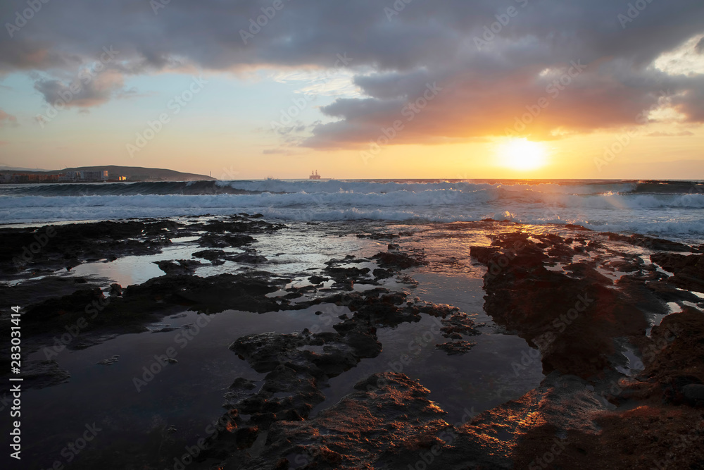Tropical sunrise over the limestone coasts of El Medano, Tenerife, Canary Islands, Spain, beautiful seascape with vibrant colors overcast sky, golden haze light and dynamic waves striking rocky shores