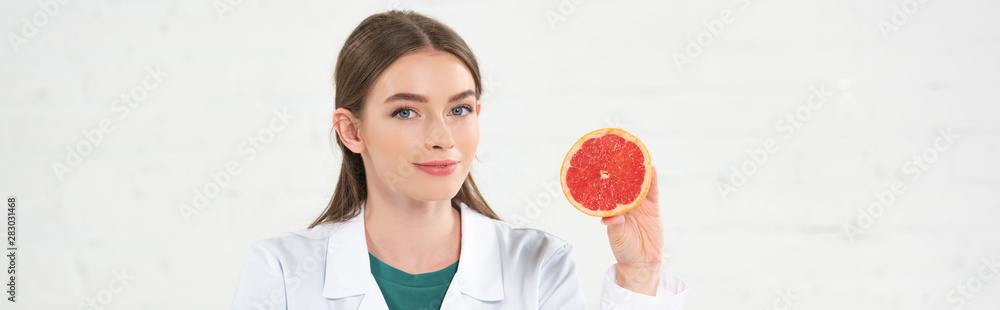 panoramic shot of smiling dietitian in white coat holding cut grapefruit and looking at camera isolated on white panoramic shot of smiling dietitian in white coat holding cut grapefruit and looking at camera isolated on white