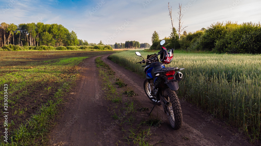 Obraz premium Tourist motorcycle with red helmet on the handlebar stands on picturesque country road, next to green field of wheat