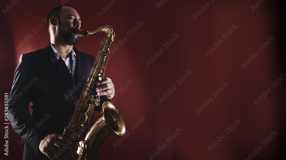 Obraz premium Portrait of professional musician saxophonist man in suit plays jazz music on saxophone, red background in a photo studio