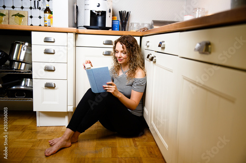 Junge Frau liest ein Buch in der Küche zu Hause. Sitzt auf dem Boden. blonde lockige Haare.