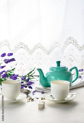 Two white tea cups with green tea  teal colored tea pot  purple flowers on white background with copy space